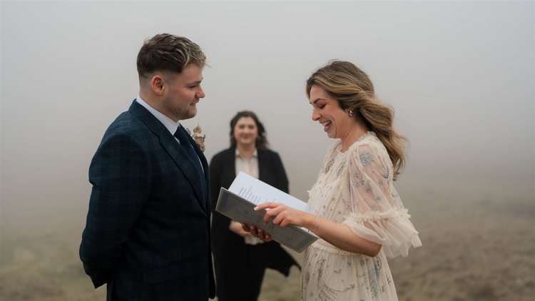 An intimate moment during a wedding ceremony set in a misty outdoor landscape. The bride and groom stand facing each other, smiling warmly - the bride holding a book or vows, seemingly mid-laughter, while the groom gazes at her affectionately. Behind