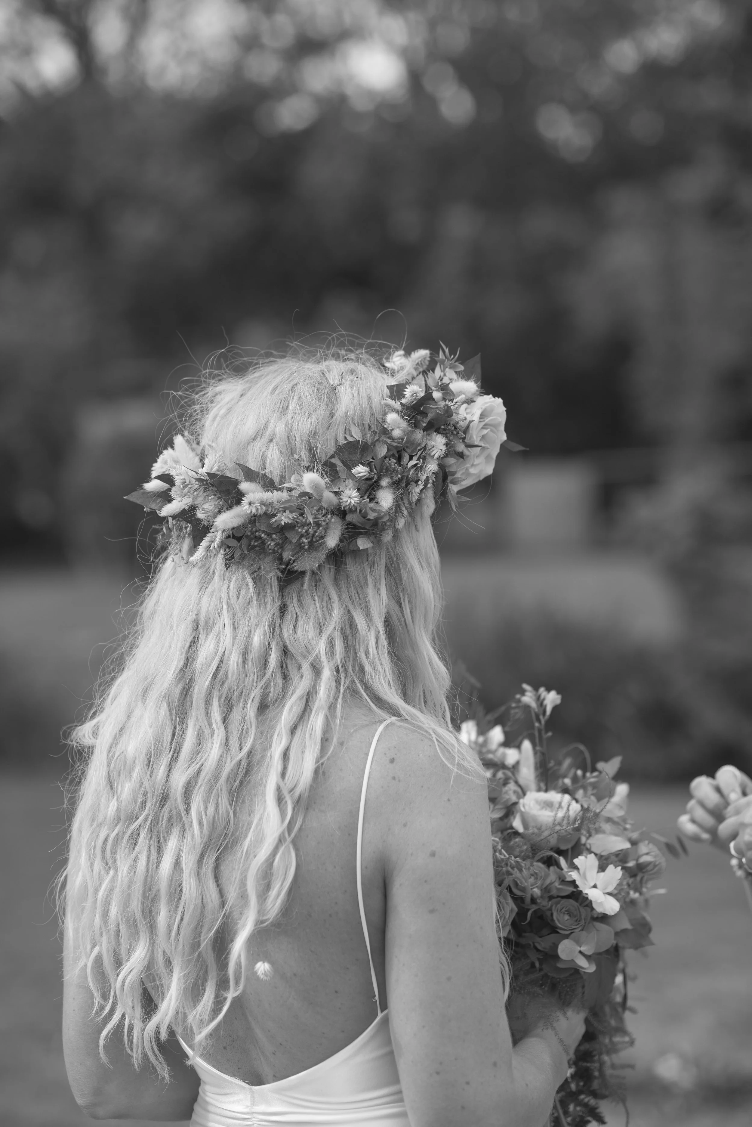 A bride with long wavy hair and a wildflower crown stands outdoors holding her bouquet, captured by Roz Pike Photography. The black and white documentary style highlights the natural textures and relaxed bohemian feel of a modern outdoor UK wedding.