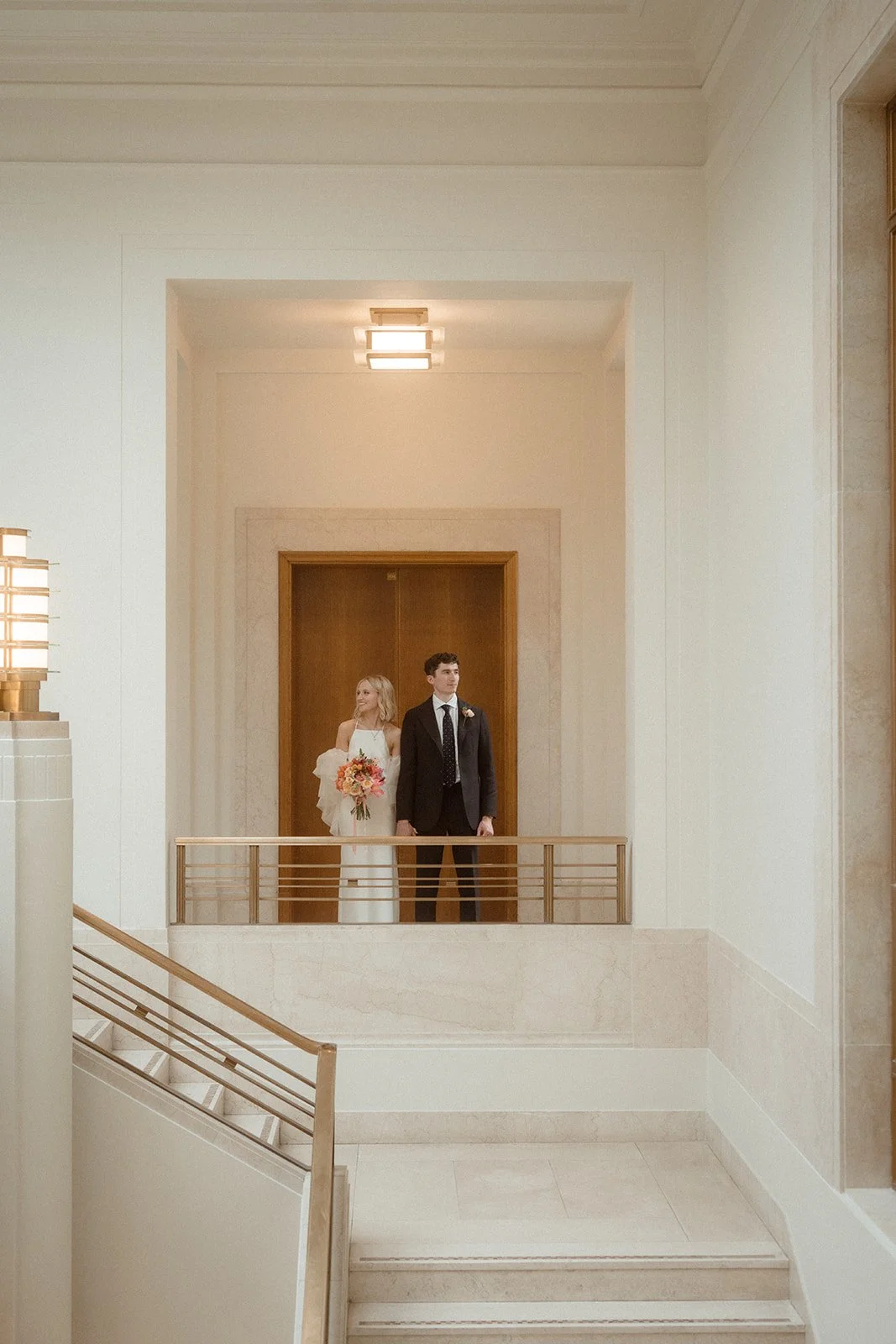 The couple stand together in a grand, softly lit interior space with tall cream walls, marble detailing and clean architectural lines while the bride holds a vibrant bouquet and the groom stands at her side; they are positioned on an upper landing be