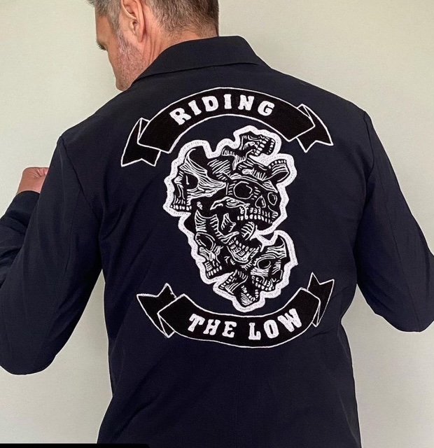 A person is shown from the back wearing a black jacket featuring a large embroidered patch with white graphic skull artwork at the centre, framed by two banner-style panels that read “RIDING” above and “THE LOW” below. The detailed stitching and bold