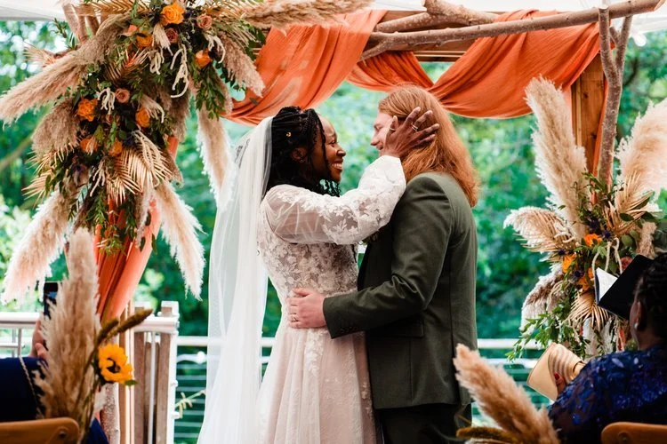 A romantic wedding ceremony styled by The Village Green Events, Sheffield. The couple share a tender moment beneath a stunning wooden arch draped in burnt orange fabric and adorned with pampas grass, sunflowers, and autumnal florals. Surrounded by lu
