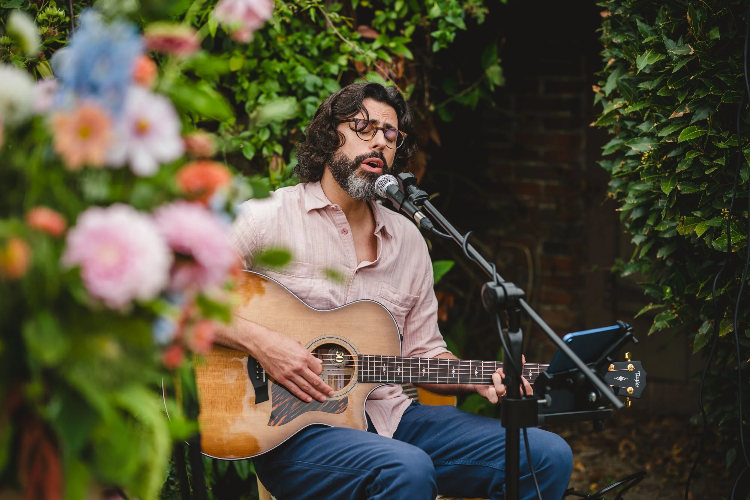 Thomas J Wilman, an acoustic wedding singer and guitarist, performs live during an intimate outdoor wedding surrounded by lush greenery and pastel flowers. His relaxed, soulful style and warm vocals create the perfect romantic atmosphere for ceremoni