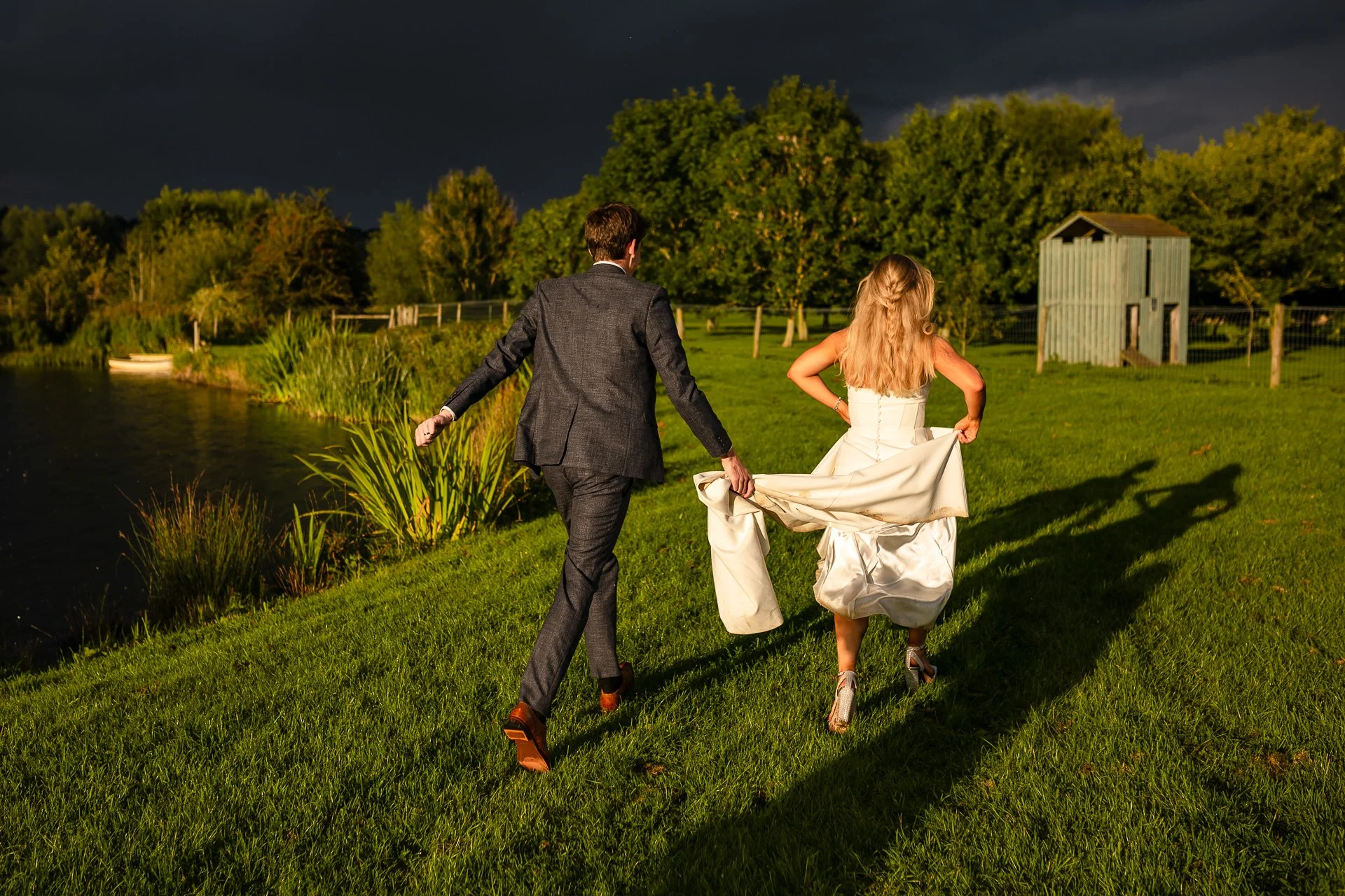 A candid wedding photo of a bride and groom running hand-in-hand across a sunlit field, the bride lifting her dress as golden hour light casts long dramatic shadows behind them. Captured by Bluebell Photo Studio, this natural and romantic outdoor wed