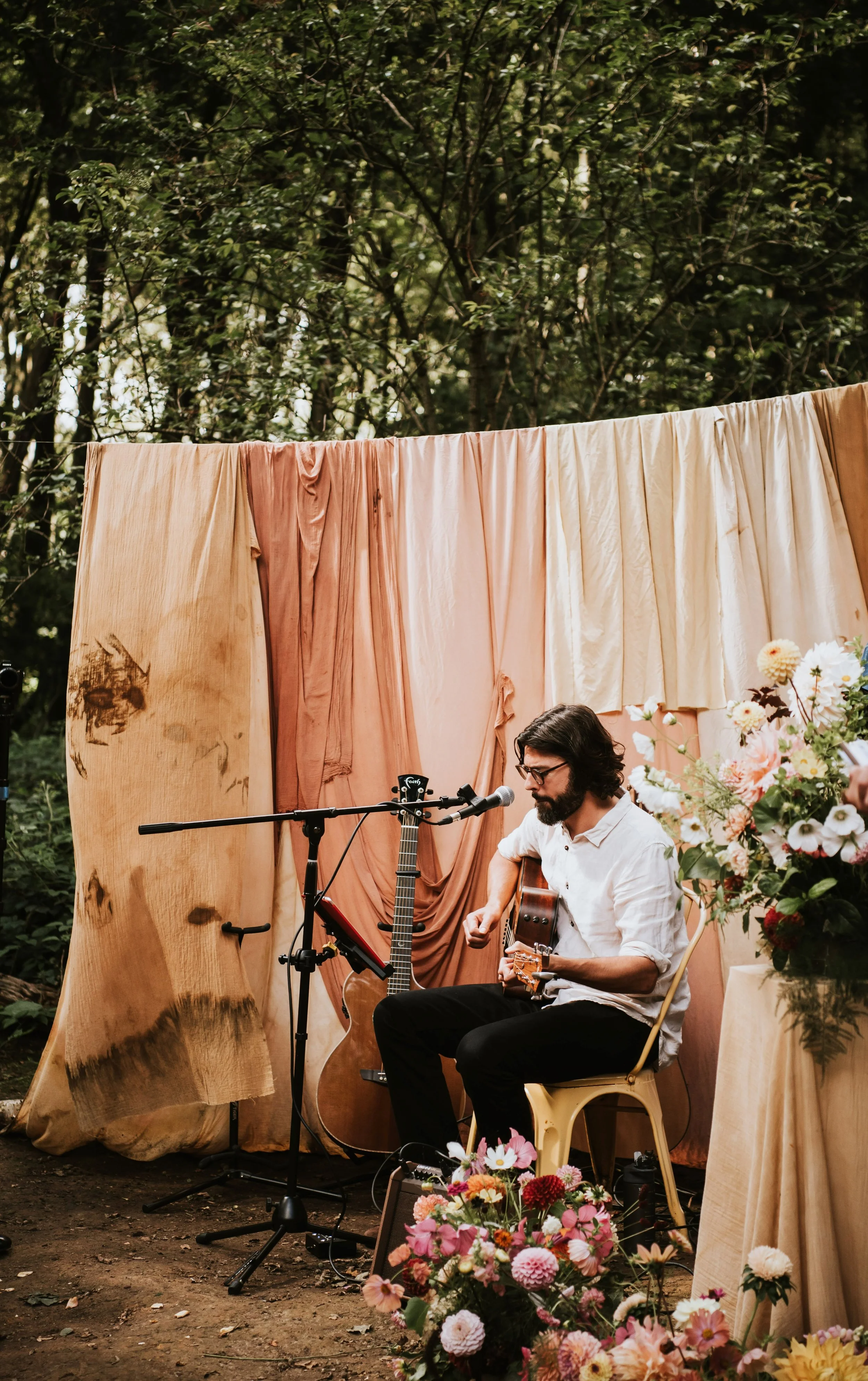 Thomas J Wilman, an acoustic wedding singer and guitarist, performs live at a beautifully styled outdoor wedding ceremony surrounded by flowers and natural woodland decor. His soulful voice and heartfelt guitar playing create a warm, romantic atmosph