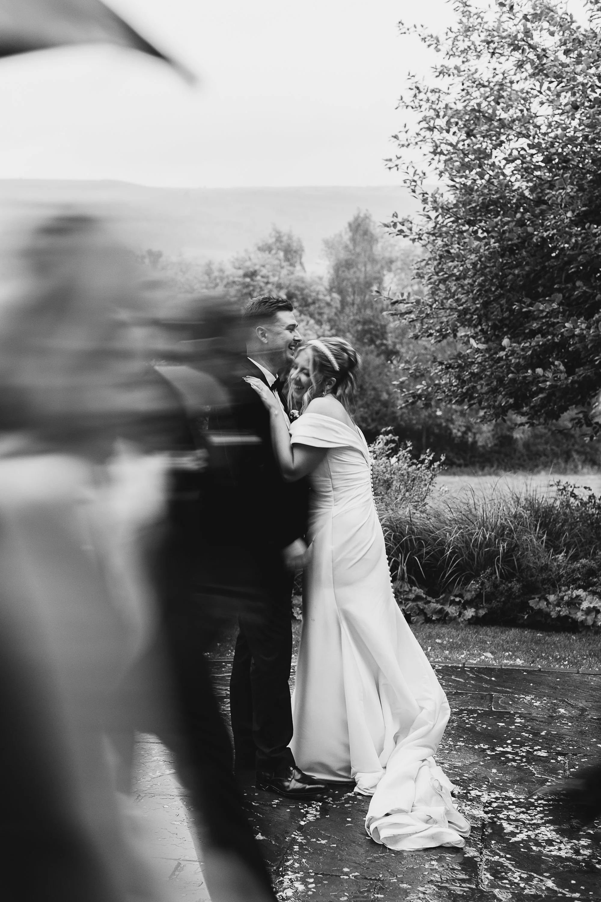 A beautifully candid and motion filled wedding moment captured by YU Photography, showing the couple laughing together as guests rush past in a blur. The black and white tones emphasise the emotion, highlighting the bride leaning into her partner wit