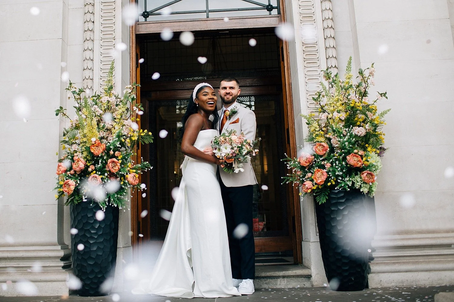 A joyful, cinematic confetti moment outside a grand stone building. At the centre, a couple stands wrapped in each other’s arms - she in a sleek, strapless white gown with a long, flowing train, he in a light beige jacket with a warm-toned tie and da