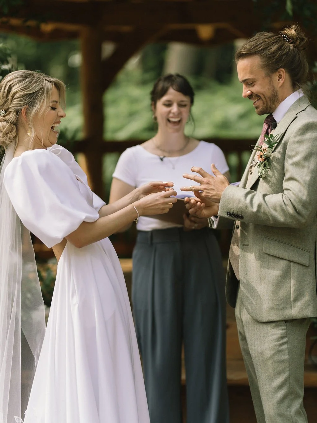 A relaxed outdoor wedding ceremony captured with laughter and love, featuring a bride in a flowing white gown and a groom in a light tweed suit exchanging rings under a rustic wooden structure. Cherry and Fern Celebrations smiles warmly as the couple