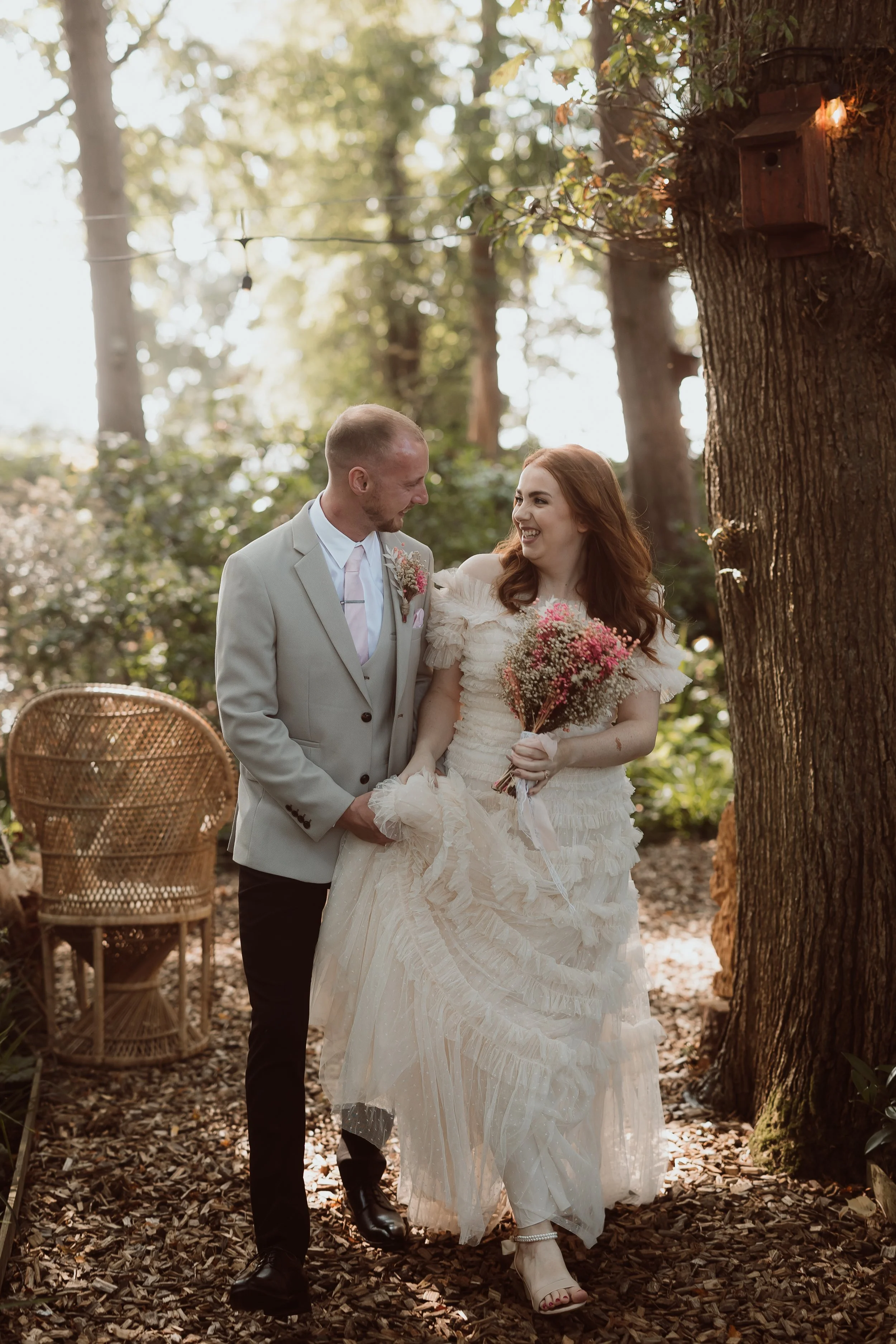 A warm and romantic woodland moment captured by Joy Louise Photography, showing the newlyweds walking side by side beneath soft golden evening light. The bride’s ruffled gown catches the sun as she lifts the hem slightly, holding a bouquet of pink an