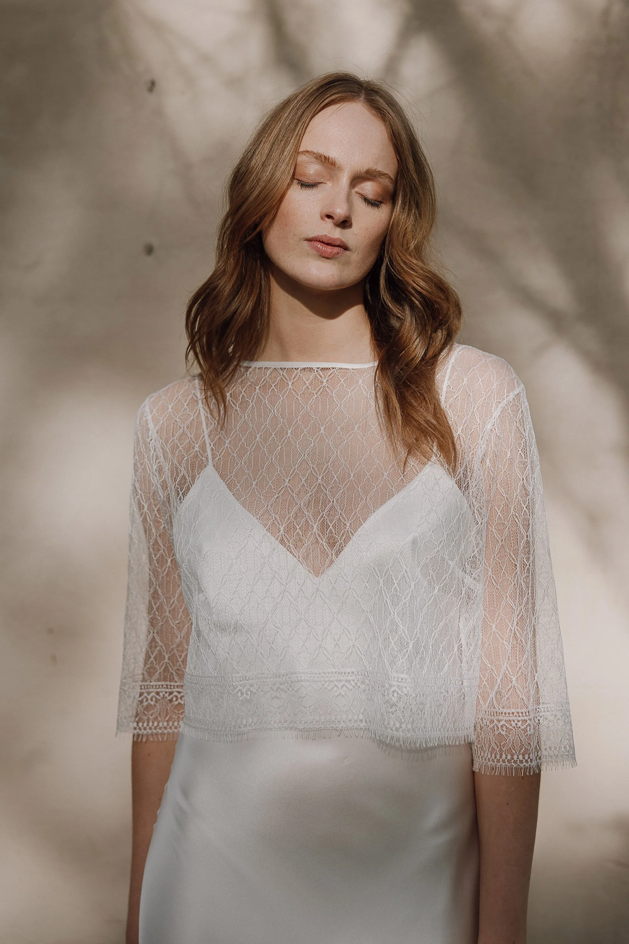 Soft sunlight filters across the bride as she stands with her eyes closed, wearing a delicate lace top layered over a smooth satin slip, the intricate pattern of the sheer fabric casting gentle shadows that echo the dappled light on the wall behind h