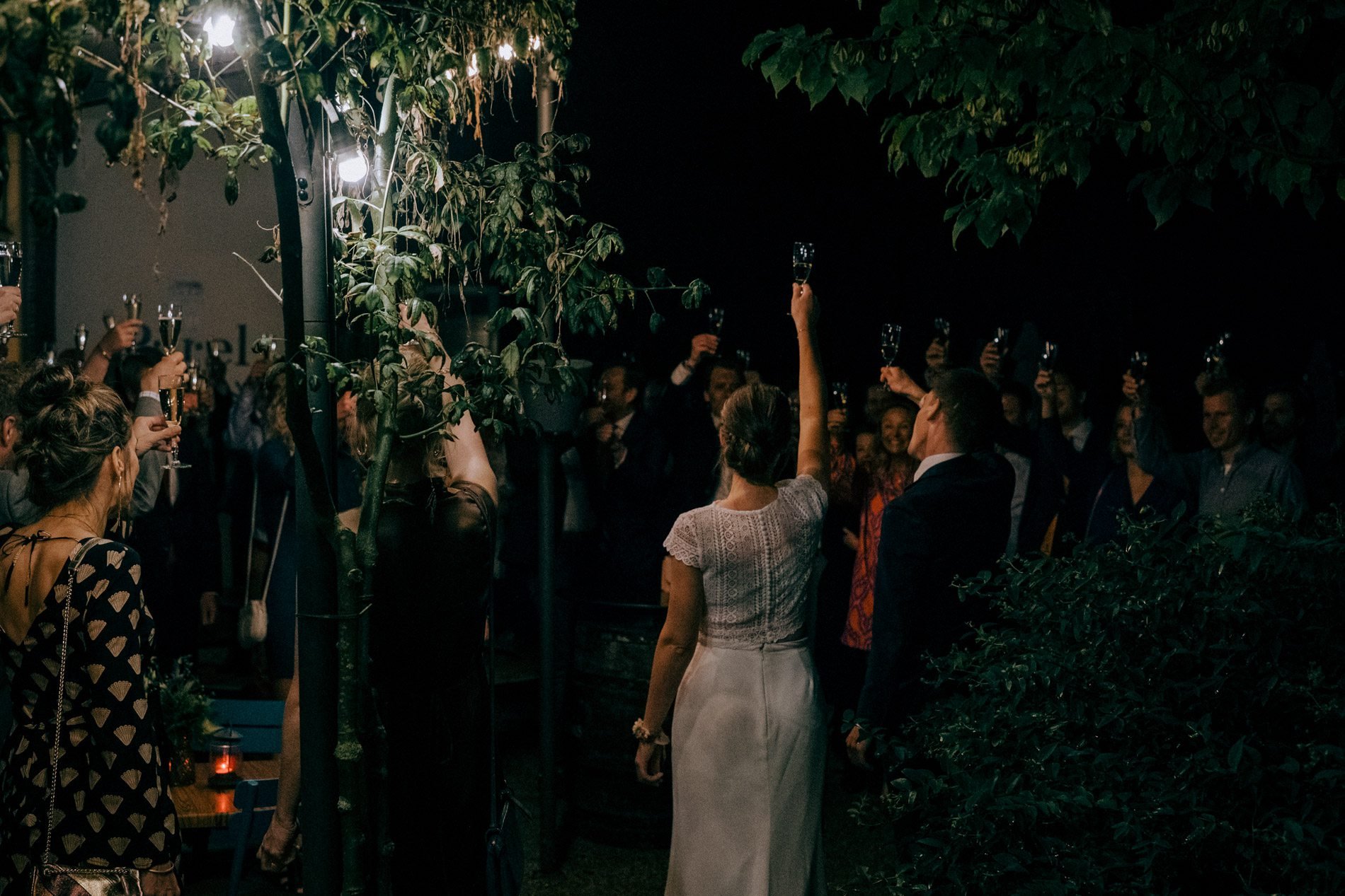 An evening wedding celebration as the couple and guests raise glasses beneath trees and lights, captured in a cinematic, documentary style by Quiet Mornings Wedding Photography in Scotland.