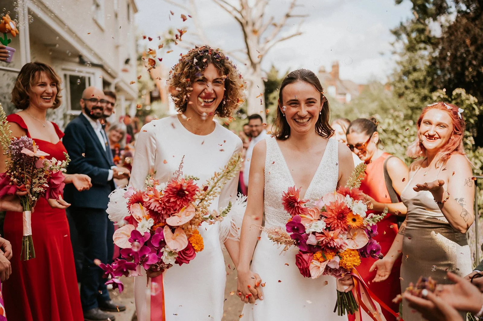 Joyful wedding moment captured by Hannah Barnes Photography, featuring two brides holding bright, modern bouquets by Harlequin Florals. The arrangements include orchids, dahlias, anthuriums, and marigolds in vivid pink, orange, and red tones, perfect