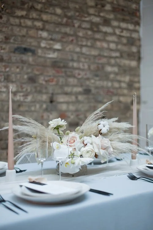 Minimal modern wedding tablescape featuring a soft neutral floral arrangement by Catherine Short Florals with blush roses, orchids, pampas grass and cotton stems. Styled with tapered candles, elegant tableware and an exposed brick backdrop, this refi