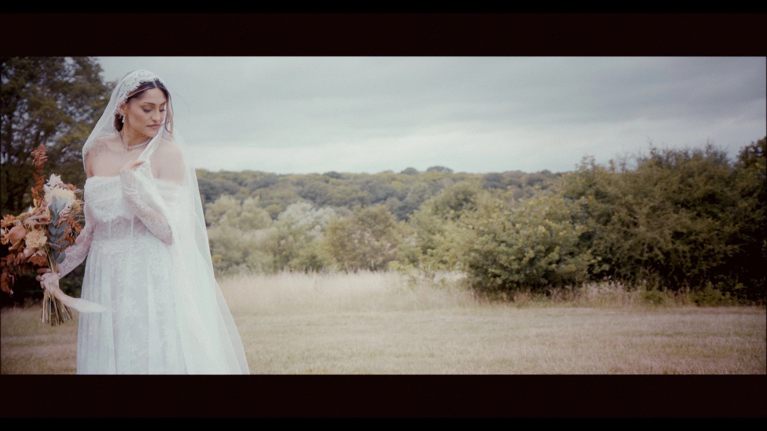 A cinematic bridal portrait by Heart Shaped Video, featuring a bride standing gracefully in a meadow under soft, overcast light. She wears an elegant lace gown with sheer sleeves and a delicate veil, holding a bouquet of warm autumnal florals. The se