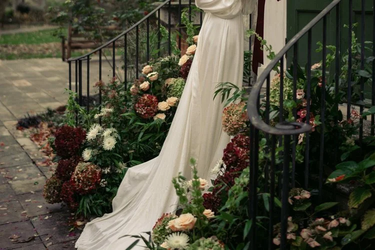 Elegant outdoor wedding floral design by Studio Clarisa Floristry featuring natural autumnal arrangements of hydrangeas, dahlias, and garden roses in warm earthy tones adorning stone steps, complementing the bride’s flowing ivory gown.