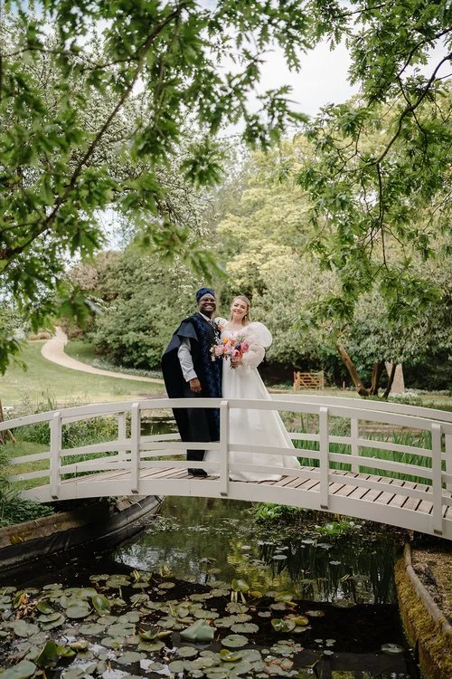 A couple stand together on a charming white bridge surrounded by lush gardens, smiling warmly as they hold colourful, seasonal flowers. The bride’s romantic gown with voluminous sleeves complements the groom’s elegant, richly textured attire, creatin