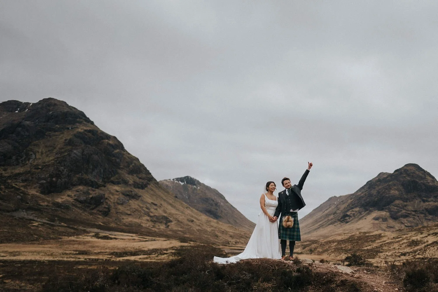 A newlywed couple stand hand in hand in a dramatic Scottish Highland landscape, with the groom in a traditional kilt raising his arm in celebration beside the bride in a flowing white dress, captured in an epic, cinematic wedding portrait by Good Luc