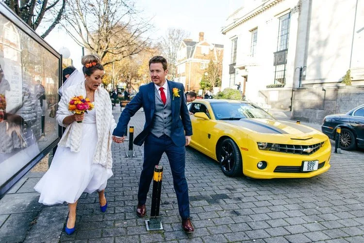 A bride and groom dash hand in hand across a city street, full of laughter and energy, with a bold yellow sports car parked behind them, captured brilliantly by Jordanna Marston Photography. The colourful flowers, sharp suits and lively urban backdro