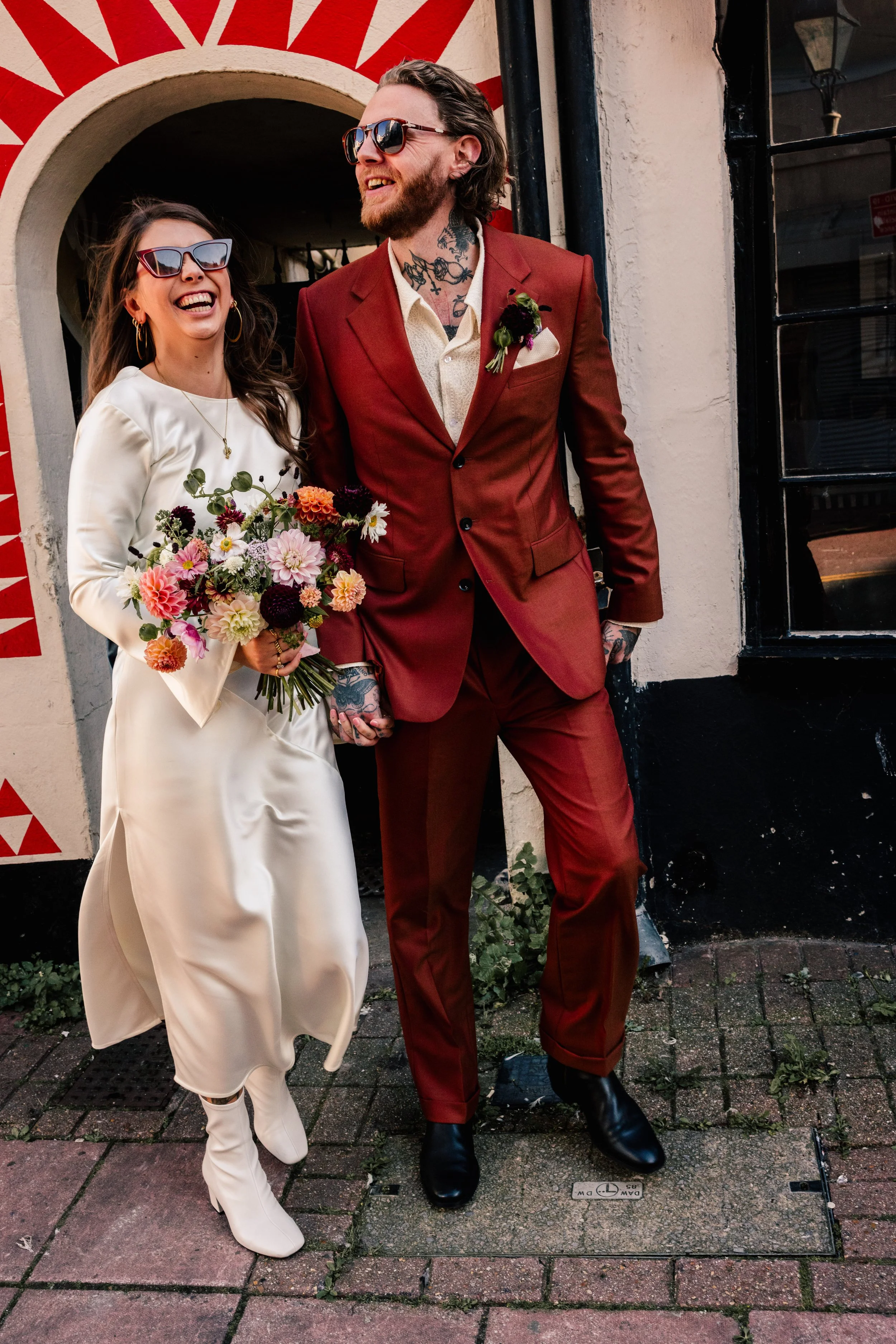 A stylish couple walks through Brighton in this vibrant wedding portrait by Teddy Photo + Film. The bride wears a chic satin midi dress with long sleeves and white heeled boots, holding a bouquet of colourful dahlias. Beside her, the groom stands out