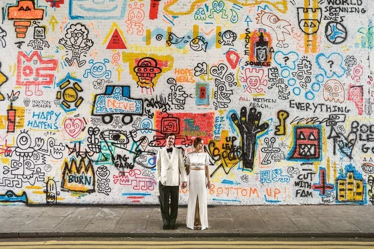 A couple stands together on a city pavement in front of a massive wall filled with bold, chaotic graffiti. The artwork behind them is a dense collage of doodles, symbols, cartoon figures, scribbled messages, and bright blocks of color. Reds, yellows,