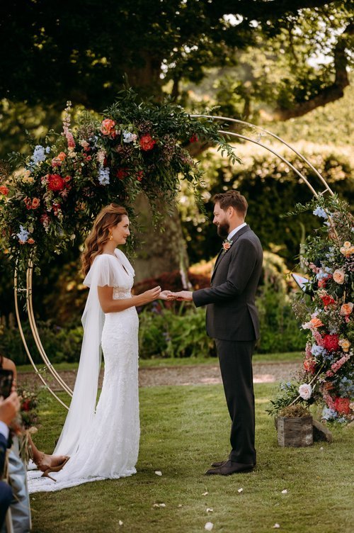 Romantic outdoor wedding ceremony featuring a floral hoop arch by The Art Bunch with vibrant dahlias, roses, hydrangeas and lush greenery arranged in a natural garden style. Framing the couple as they exchange vows on a sunlit lawn, this colourful ar