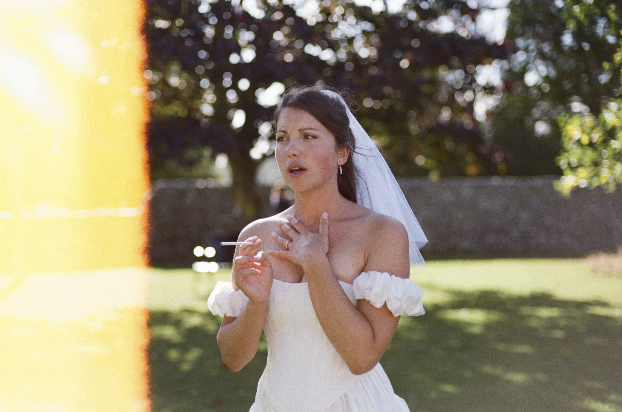 A bride stands in soft afternoon light with a veil and off-the-shoulder gown, captured by Roz Pike Photography. Shot on film with a warm light leak, the candid moment captures raw emotion and relaxed documentary wedding style in a sunlit garden setti