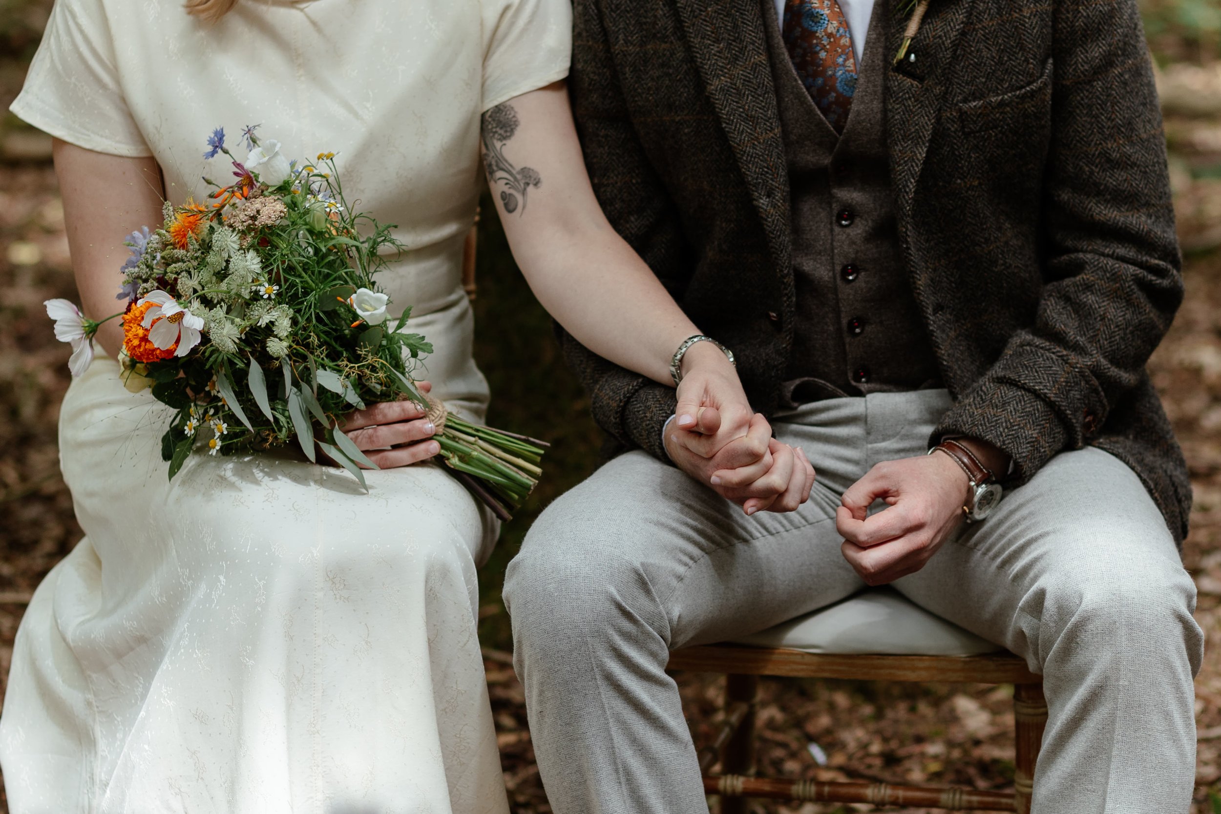 A cool photo of a modern bride and groom holding hands at their woodland wedding ceremony. The bride is holding a wild flower bouquet and their heads are cropped out.