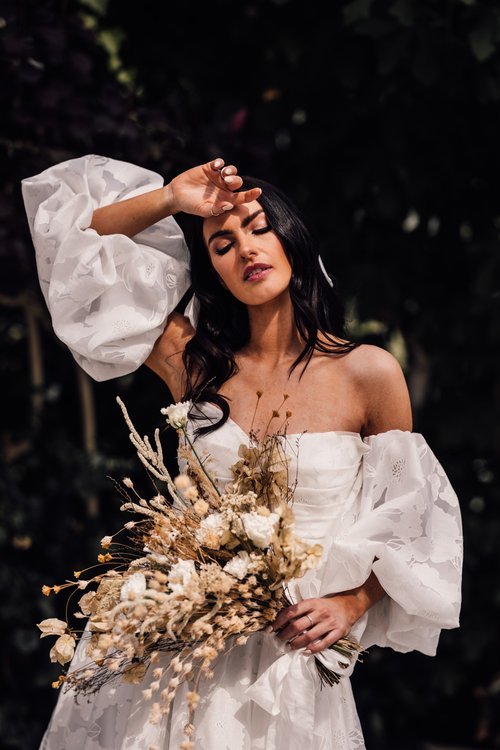 Romantic bridal portrait featuring soft glam makeup by Glamorama Make-up Artist Liverpool. The bride wears a modern off-shoulder gown with statement sleeves and holds a bouquet of dried flowers, creating a dreamy, natural look ideal for contemporary 