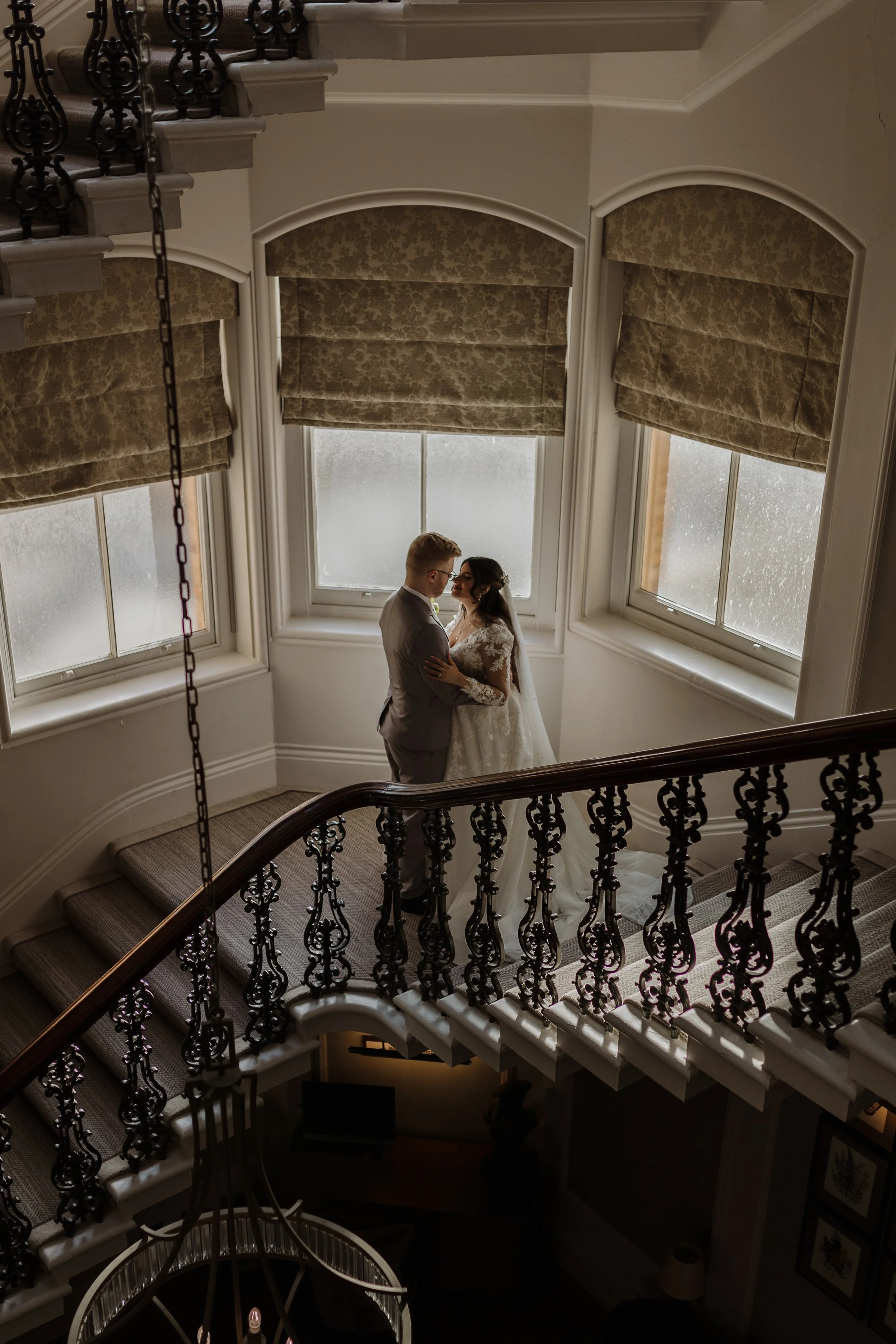 A beautifully atmospheric moment curated by Big Sister Events, featuring a couple sharing an intimate embrace on an elegant sweeping staircase framed by ornate ironwork and soft natural window light creating a timeless romantic aesthetic perfect for 