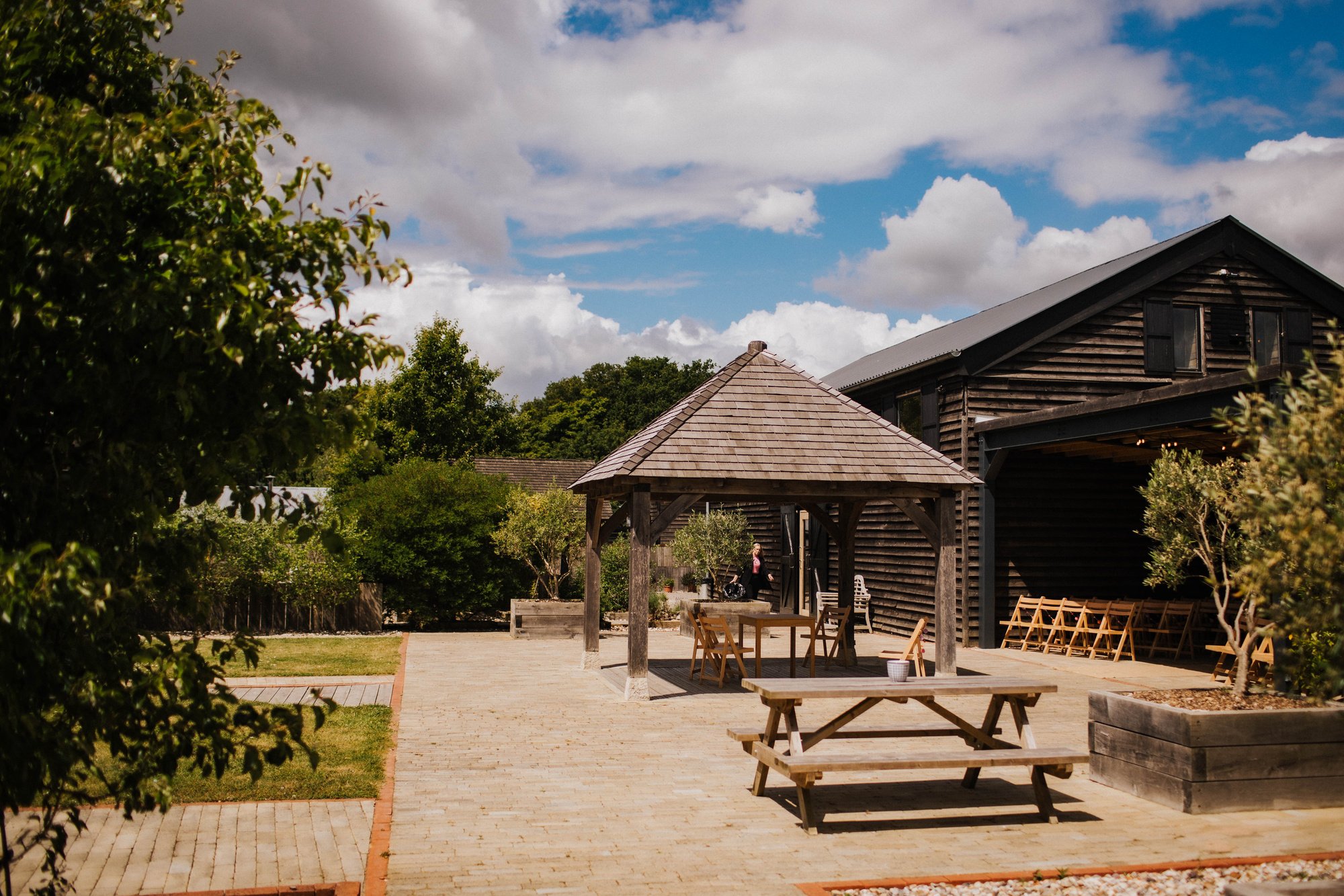 A sunlit courtyard features a timber gazebo with a shingled roof at its center, surrounded by planted borders, neat paving and wooden seating, while the dark clad barn of The Canary Shed opens onto the space and leafy trees frame the view beneath a s