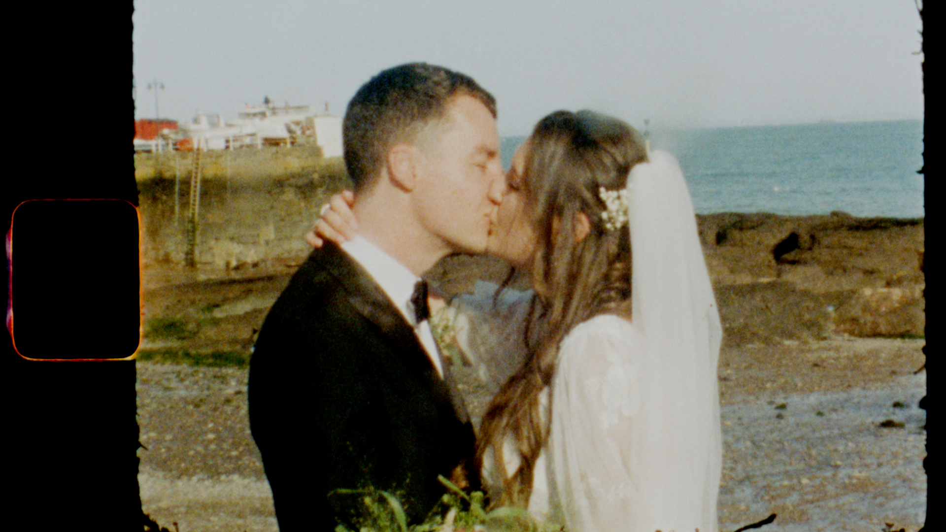 A romantic still of a bride and groom kissing by the sea. It has been taken using Super 8 film.