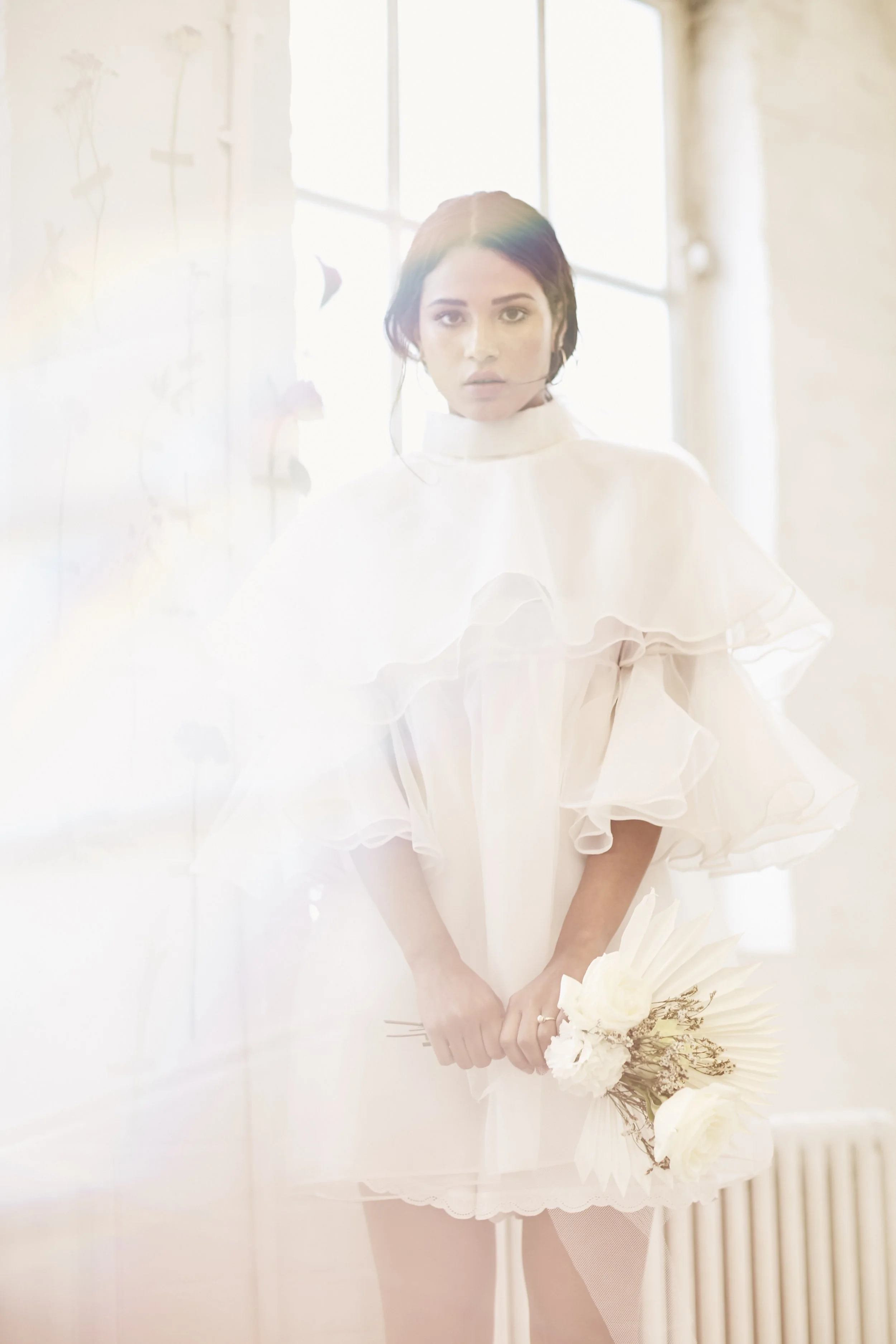 Soft light floods the frame, partially veiling the figure and blurring the edges of the scene. Layered sheer fabric forms gentle ruffles around the shoulders and bodice, creating volume without weight. The short silhouette is balanced by transparency