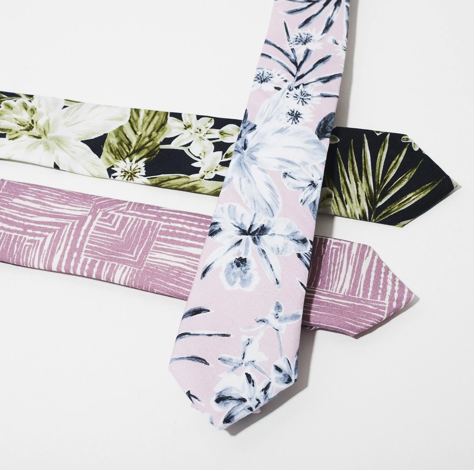 Three patterned Francis + Louise ties laid out on a white surface, each featuring a different design and colour palette. The tie on top has a soft pink background with large white and grey floral illustrations, the tie beneath it has a geometric patt