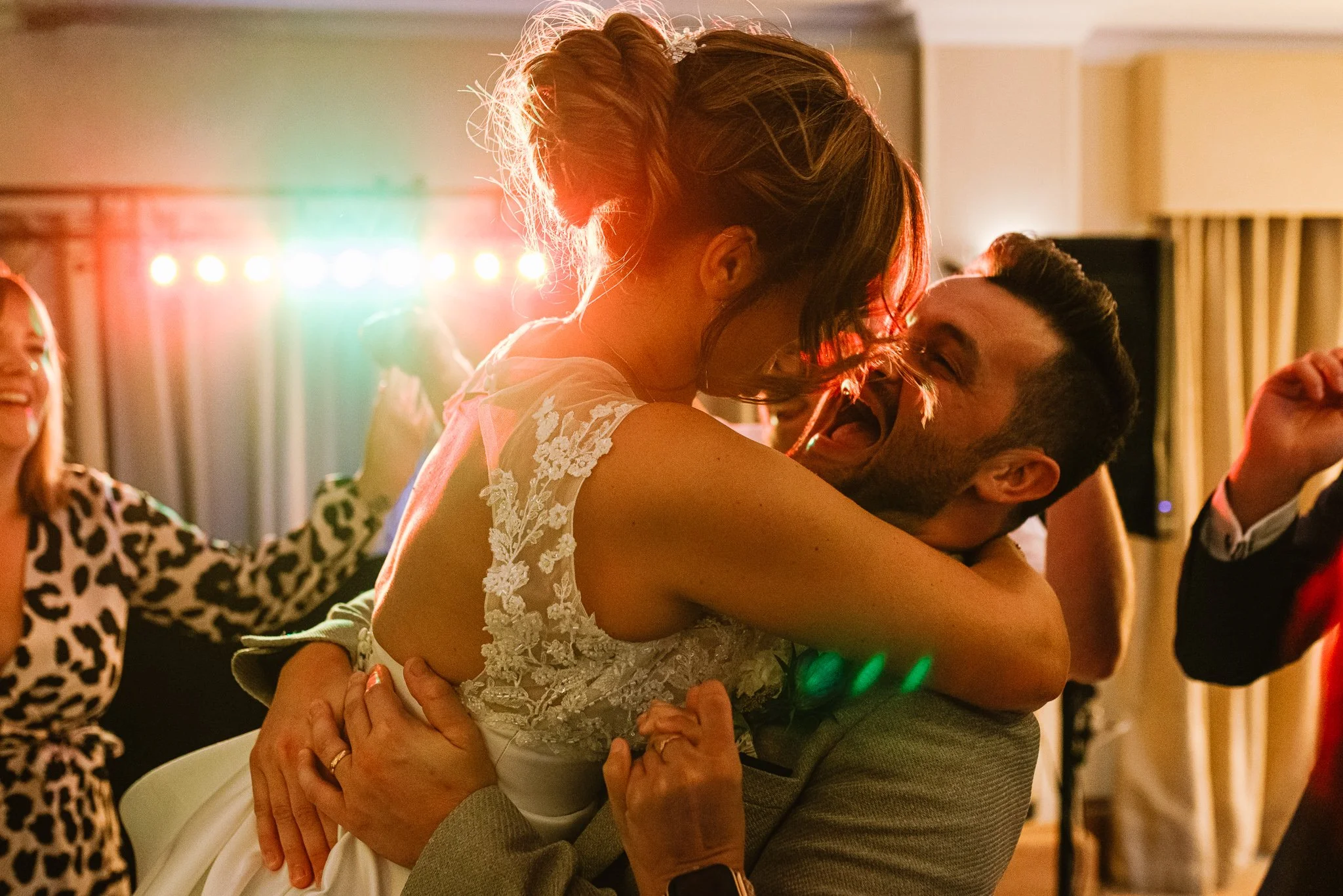 A joyful wedding reception moment captured by Bluebell Photo Studio, showing the bride being lifted into her partner’s arms on the dancefloor as colourful party lights glow behind them. The couple laugh together in a candid, emotional embrace while g