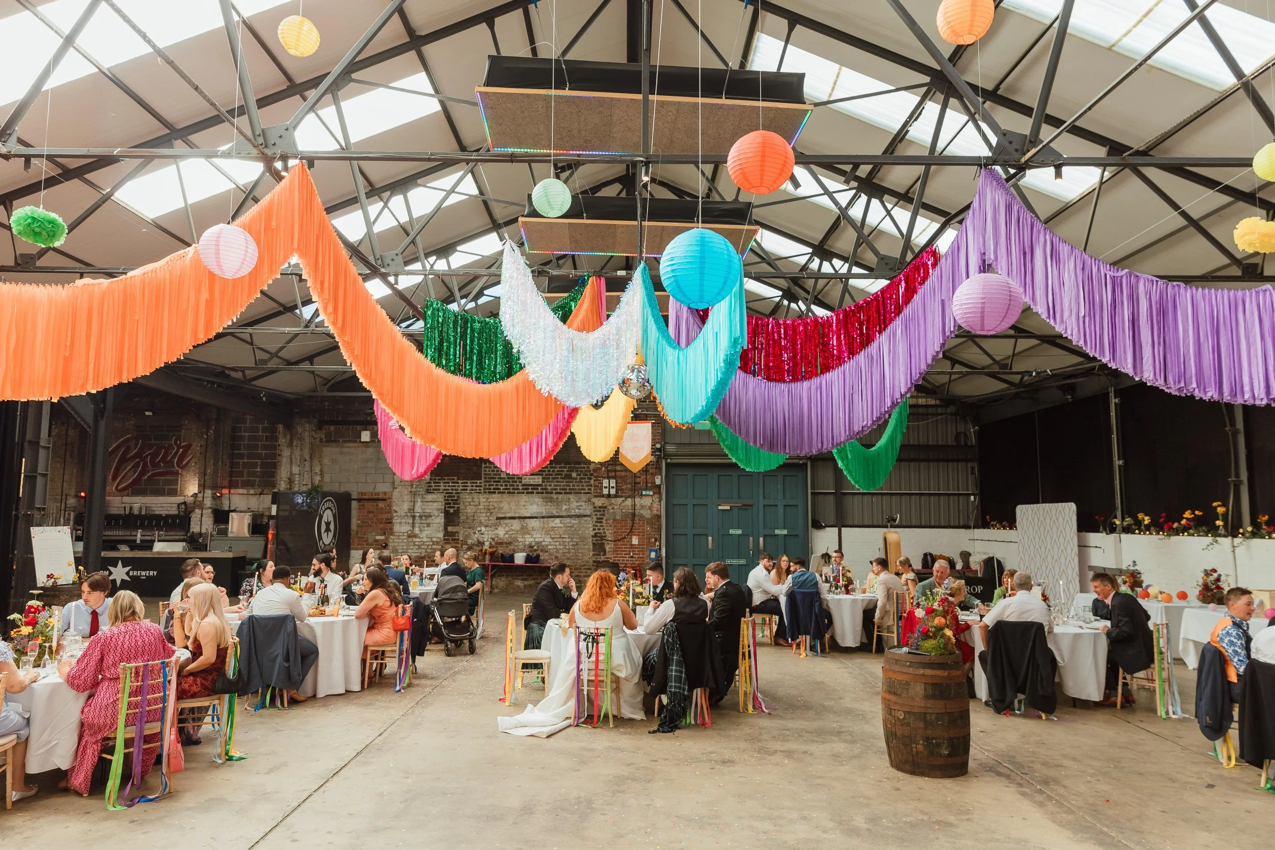 Colourful Streamadelica wedding reception inside an industrial warehouse venue, featuring vibrant fabric draping, hanging lanterns and long banquet tables for a creative, festival-style celebration.