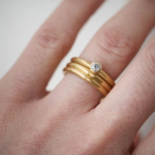 A minimalist Natalie Vernazza diamond solitaire engagement ring stacked with slim yellow gold wedding bands, featuring a small bezel-set round diamond for a modern, understated bridal jewellery look.