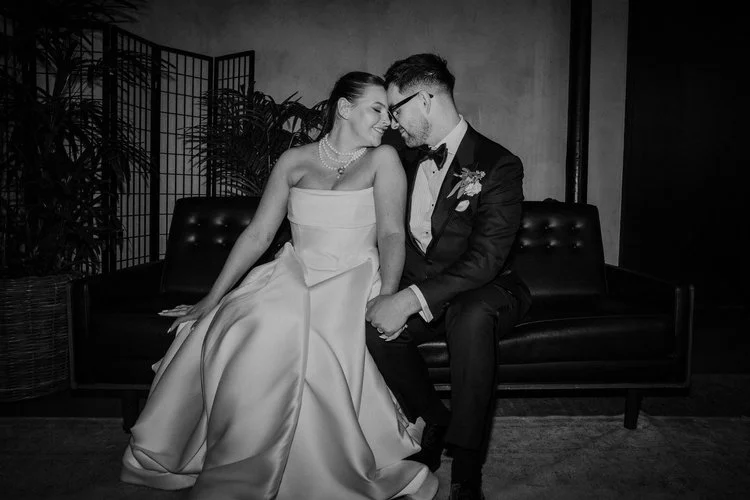 A sophisticated black-and-white wedding portrait captured by Mirrorbox Photography, showing a couple seated closely together on a modern leather sofa. The bride’s structured, strapless gown drapes beautifully around her as she leans in toward her par