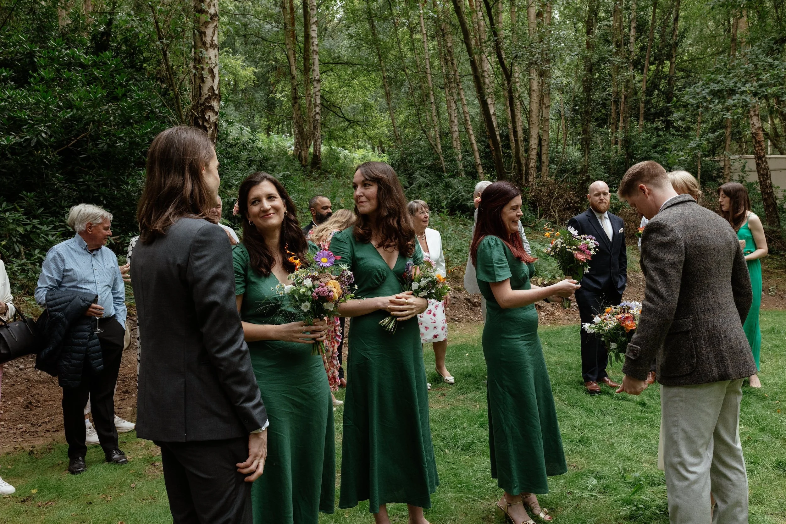 Bridesmaids wearing green dresses and chatting to other guests at a woodland wedding. They are holding colourful bouquets.