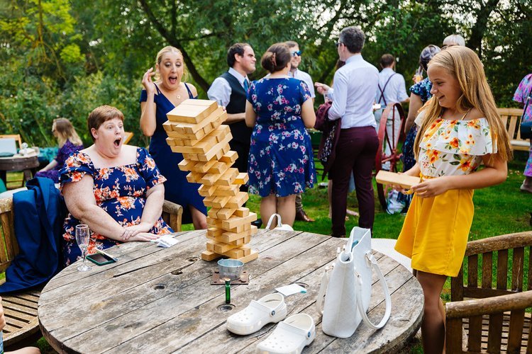 Wedding guests playing a giant outdoor Jenga game during a summer wedding reception. A young girl pulls a wooden block as the tower dramatically tilts, while two women react with shock and laughter. Other guests in floral outfits chat in the backgrou