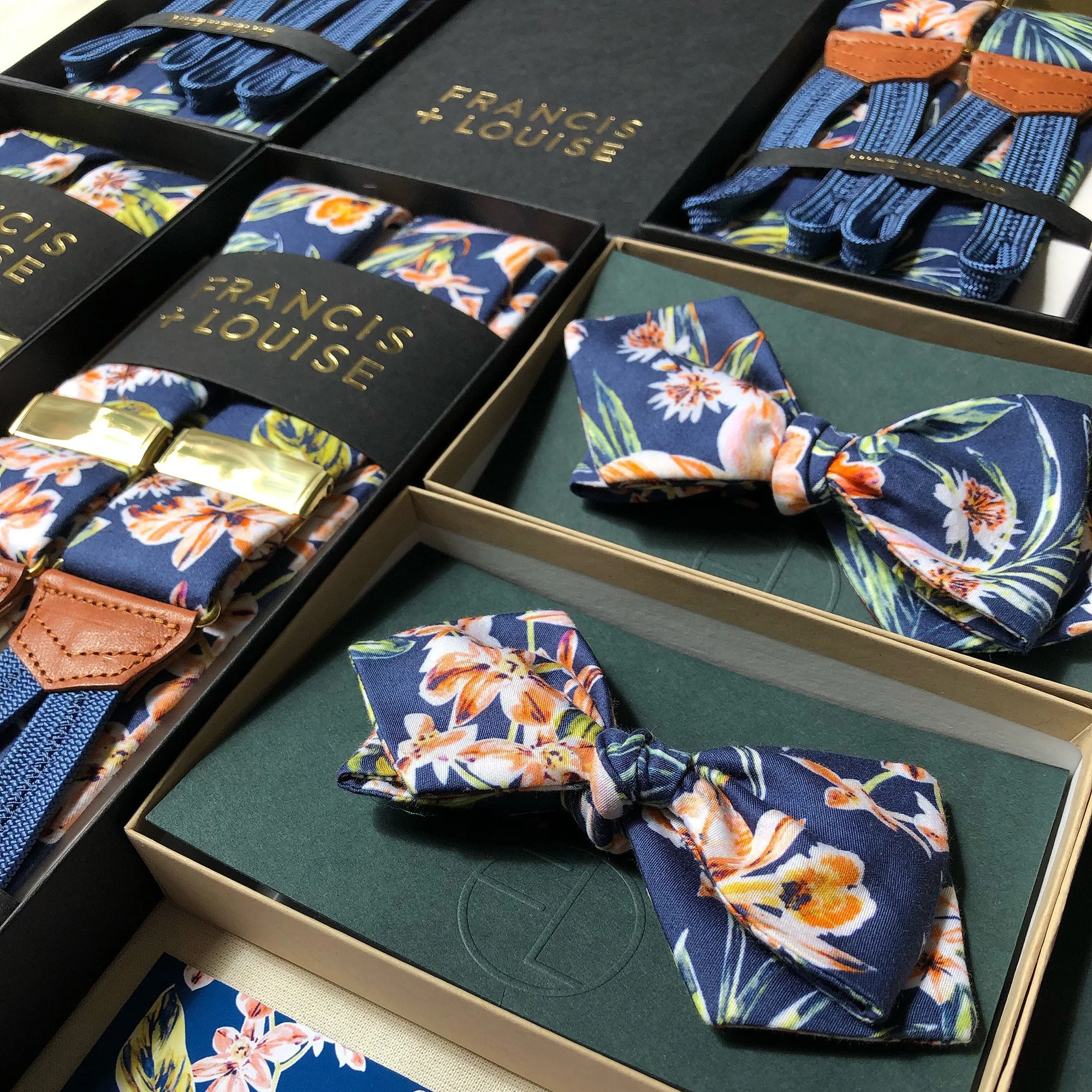 A collection of Francis + Louise accessories arranged in their boxes, featuring suspenders and bow ties in a bold navy floral print with orange, white and green accents. The suspenders are presented with gold clips and tan leather detailing, while th