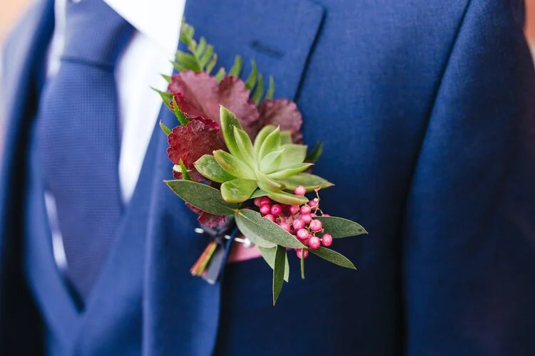 Modern groom’s buttonhole by Catherine Short Florals featuring a fresh succulent, pink hypericum berries, heuchera leaves and soft greenery. Styled against a sharp navy suit and textured tie, this contemporary natural design highlights Catherine Shor