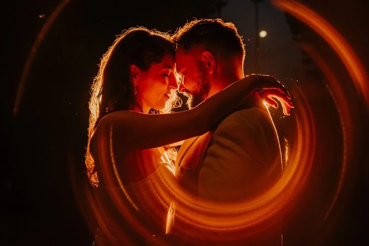 A couple share an intimate moment during their evening wedding celebration, illuminated by warm creative lighting that forms a glowing circular frame around them, captured by Joshua Humphrey Photography. The dramatic backlight and close embrace creat