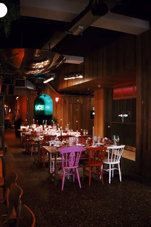 A stylish wedding reception setup inside Pier Court  Brighton, featuring a mix of colorful chairs, rustic wooden tables, and soft candlelight. The modern industrial interior combines wood paneling, neon lighting, and a relaxed atmosphere, creating th