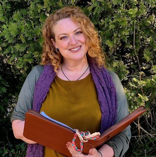 Melly the Celebrant standing outdoors holding a leather ceremony folder, surrounded by lush greenery. She wears an olive green top with a purple scarf, radiating warmth and friendliness. A natural, welcoming portrait perfect for showcasing a professi
