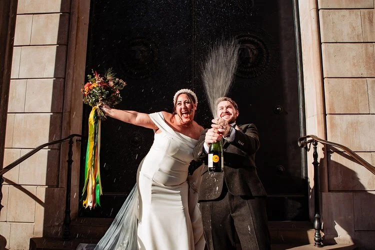 A lively and joy filled wedding moment captured by Natalia Case Photography, showing the newly married couple celebrating on the steps outside their venue. The bride’s bright expression and raised bouquet create an instant sense of excitement, while 