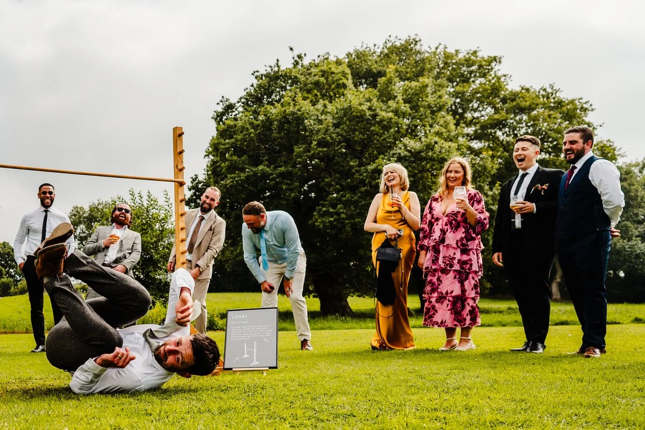 A group of guests burst into laughter on a sunny lawn as one man dramatically rolls on the grass beneath a limbo pole, clutching a drink while his friends double over watching, their bright outfits and carefree expressions giving the whole scene a pl