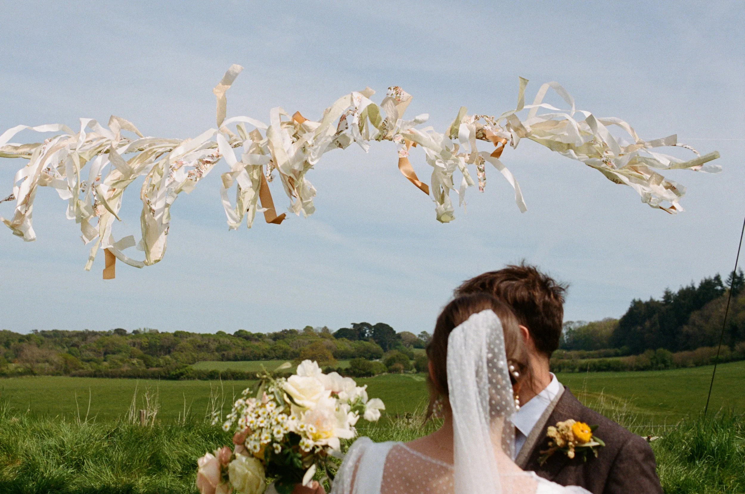 A couple stand together beneath flowing ribbon decorations in a wide open countryside setting, captured by Roz Pike Photography. The bride’s dotted veil and soft pastel bouquet complement the natural rural landscape, creating a dreamy romantic moment