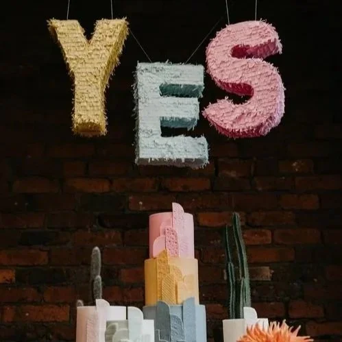 A playful wedding display featuring pastel piñatas spelling 'YES' hanging above a colorful cactus-themed cake and vibrant floral accents, created by Pinyatay. Photo by Nicki Shea Photography.