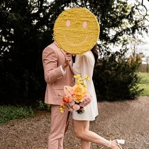 A playful wedding portrait of a couple kissing behind a large yellow smiley face piñata, with the bride holding a vibrant bouquet, created by Pinyatay. Photo by Stephanie Butt Photography.