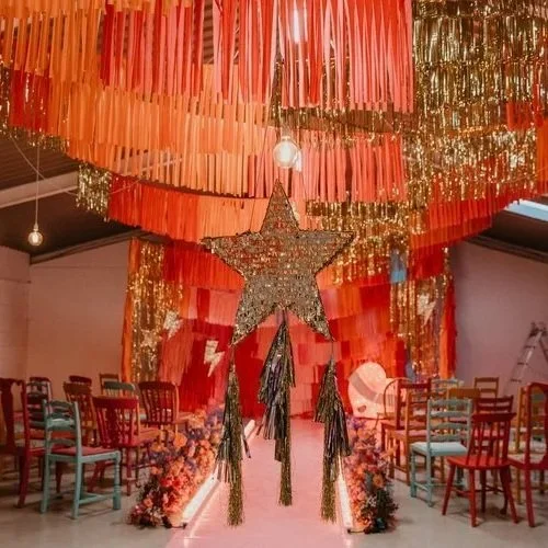 A bold and colorful indoor wedding setup with a large glittering star piñata, hanging fringe garlands in red and gold, and vibrant mismatched chairs, created by Pinyatay.
