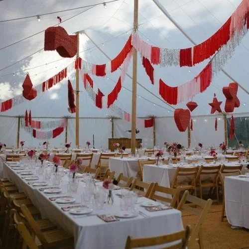 A festive wedding reception under a white tent decorated with red and white fringe garlands and hanging piñatas in various shapes, with long banquet tables set for guests, created by Pinyatay.